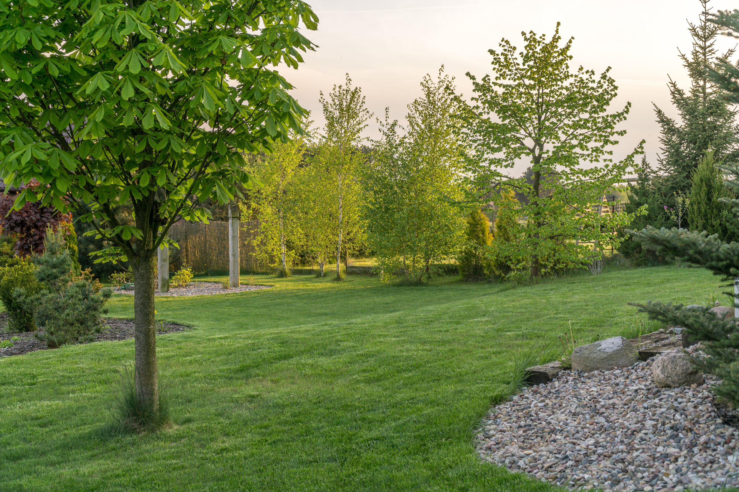 A well-maintained garden with lush green grass, several leafy trees, and neatly arranged rocks on the right side. The scene is bright and peaceful, with sunlight filtering through the trees.