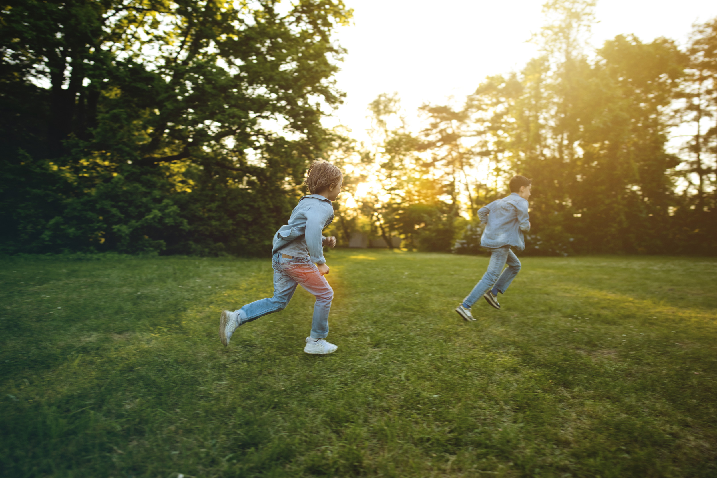 Two children wearing denim jackets and jeans run across a grassy field in a park, surrounded by green trees, with sunlight streaming through the branches in the background.