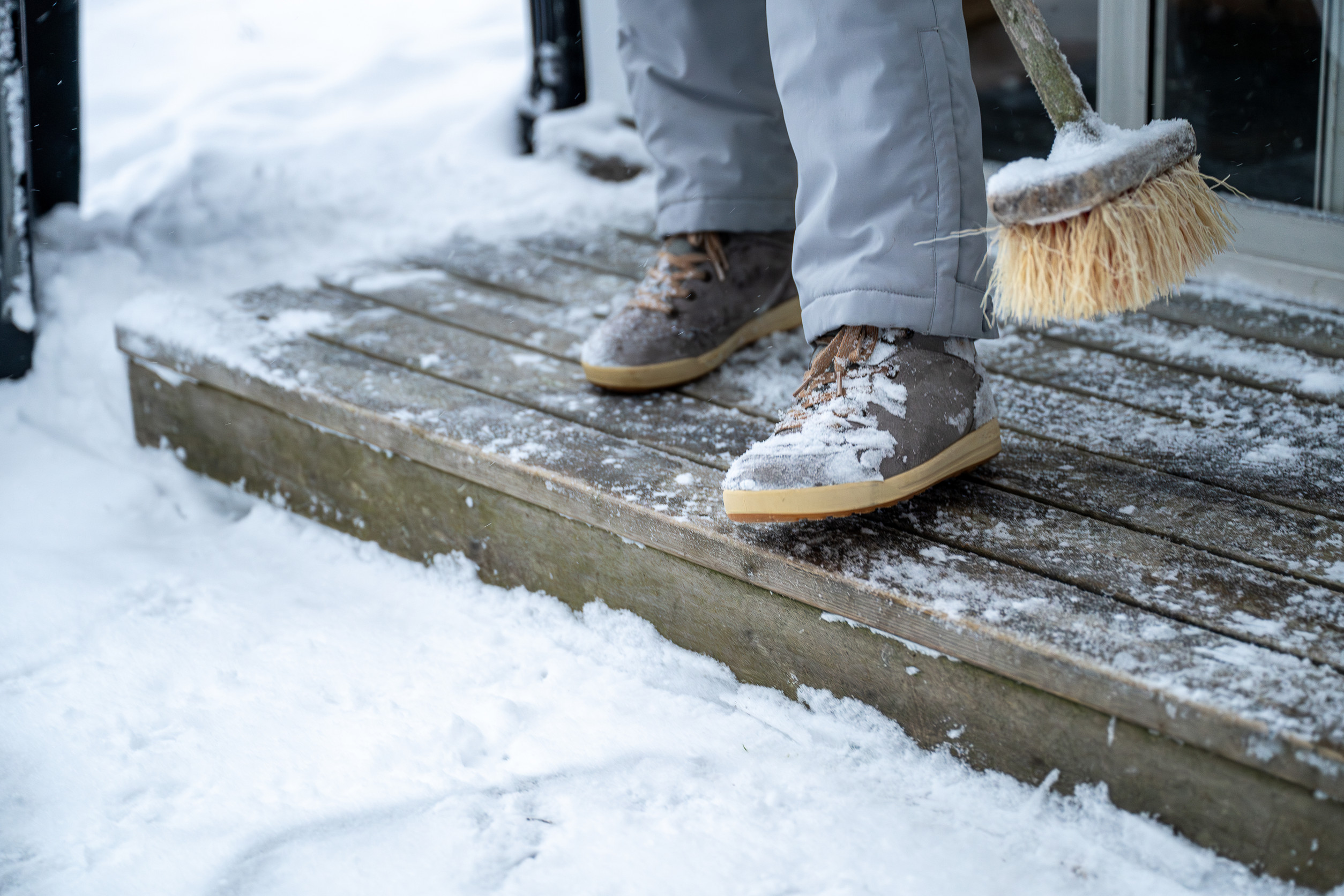 A person wearing winter boots and gray pants sweeps snow off a wooden porch with a broom. Snow covers the ground and porch edges, and the scene appears cold and wintry.