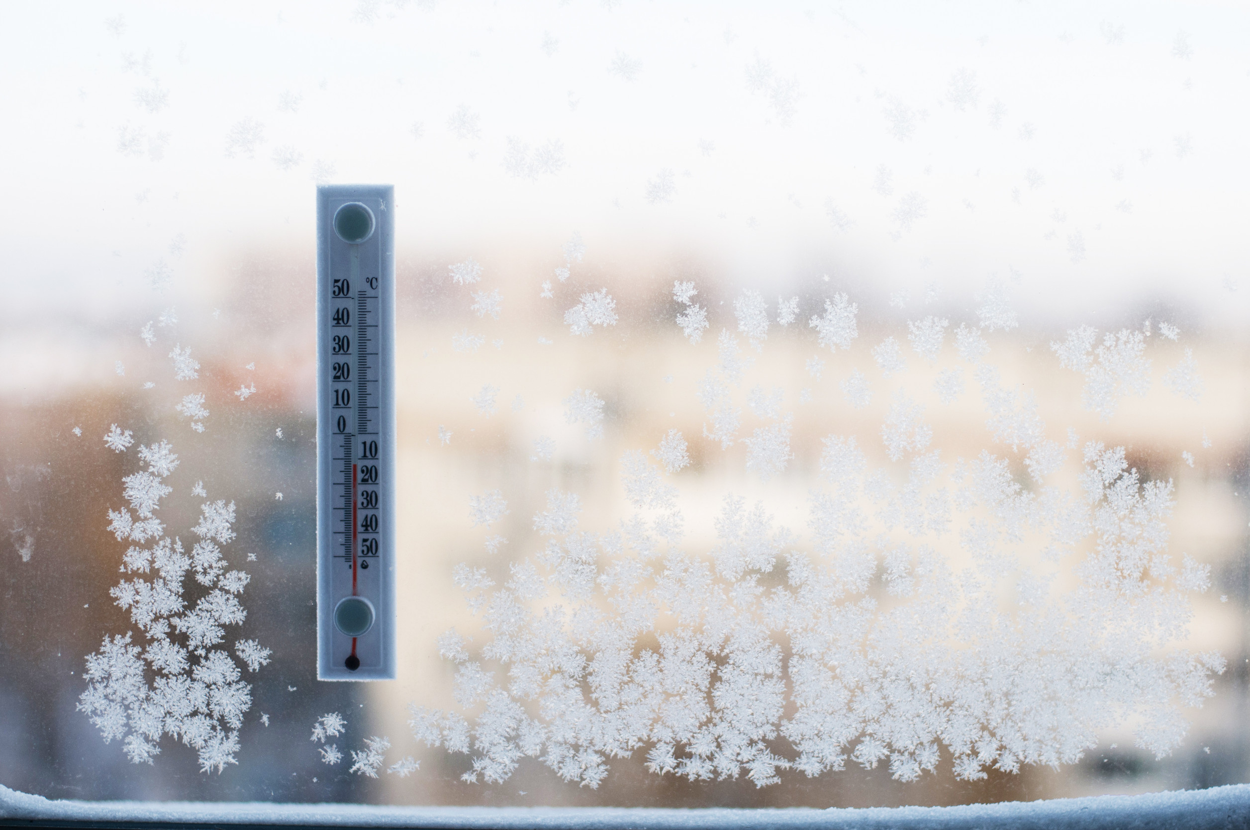 A thermometer attached to a frosted window shows a temperature below zero Celsius, with intricate ice crystals covering much of the glass and a blurred outdoor scene in the background.